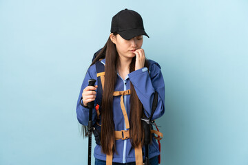 Young Chinese girl with backpack and trekking poles over isolated blue background having doubts