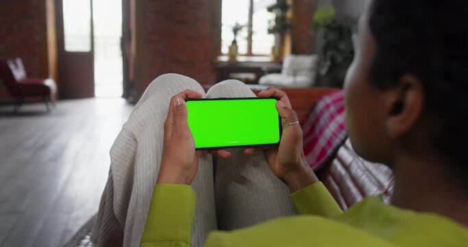 Young woman lying on couch with modern smartphone in his hand while looking on mock up screen with trecking markers on it. Concept of green screen and chroma key. Cosy home background