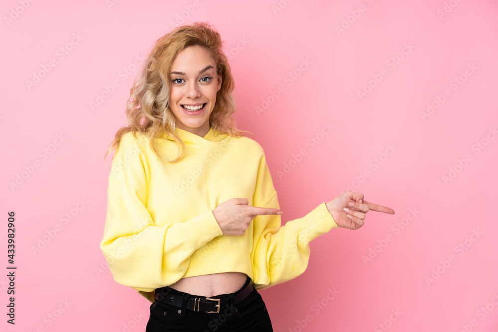 Poster Young blonde woman wearing a sweatshirt isolated on pink background surprised and pointing side