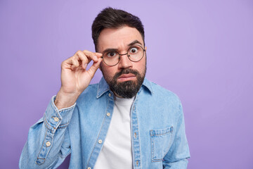 Indoor shot of handsome surprised adult man with thick beard looks attentively through round spectacles dressed in denim shirt hears exciting news stands speechless against purple background