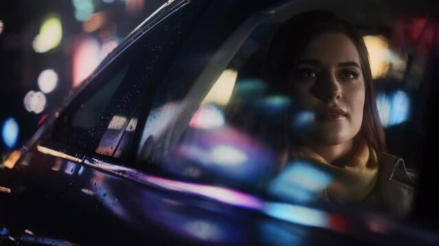 Stylish Female is Commuting Home in a Backseat of a Taxi at Night. Beautiful Woman Passenger Looking Out of Window while in a Car in Urban City Street with Working Neon Signs. Cinematic Footage.