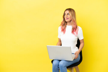 Naklejka premium Young woman sitting on a chair with laptop over isolated yellow background and looking up