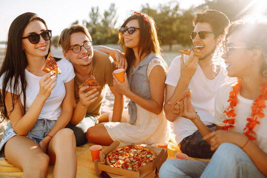 Group Of Young Friends Having Picnic, Eating Pizza, Toasting With Beerus At The Beach. Happy Friends Resting Together Sitting Near The Sea. Fast Food Concept. Beach Holiday And Summer Vacation Concept