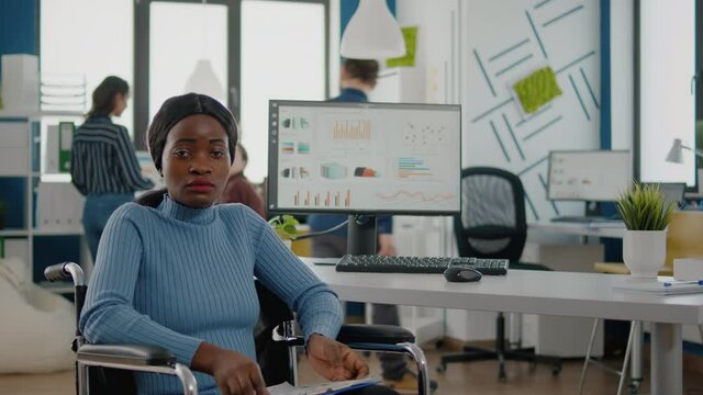 Young black woman with disabilities, invalid, disabled paralized handicapped looking at camera frustred, sad sitting in wheelchair in business office room, working at financial project with team.