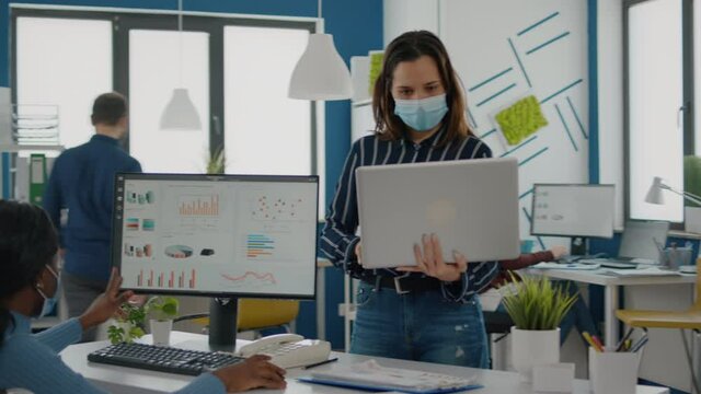 Company manager with mask against coronavirus explaining to african paralysed colleague project plan standing near desk holding laptop. Multiethnic business team working respecting social distance.