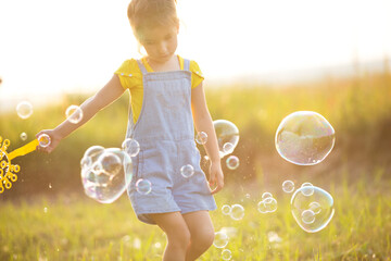A girl in a denim jumpsuit blows soap bubbles in the summer in a field at sunset. Children's Day, happy child, outdoor activities in the summer. Summer background. Healthy and eco-friendly lifestyle