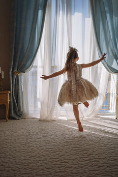 Back view of little girl in white dance dress dancing by the window in luxury apartment bedroom and looking out at the sea. Image with selective focus
