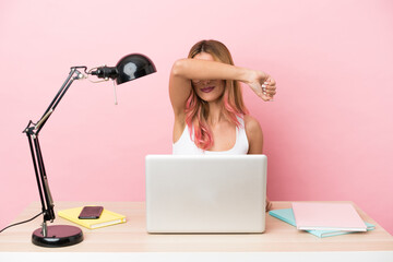 Young student woman in a workplace with a laptop over pink background covering eyes by hands