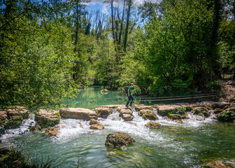 Adventurous blonde Caucasian woman crossing a river jumping on boulders by a small waterfall in the middle of a lush green forest in summer. Sentierelsa, Colle di Val d'Elsa, Siena - Tuscany, Italy