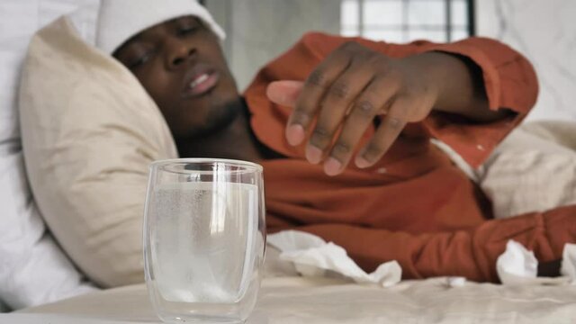 African-American man with patch on head suffering from cold puts soluble tablets into glass with water lying on bed close view