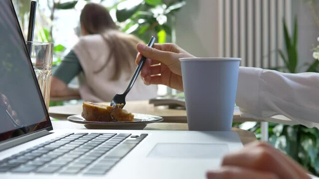 Woman with coffee and laptop takes piece of delicious cake using fork at table in cafe extreme close side view slow motion