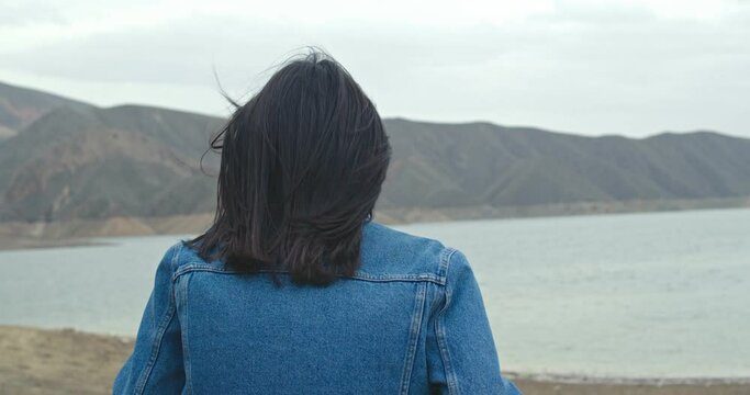Close back view of  woman hair tender movement in the air in slow motion.  Woman in jeans jacket  walking on sandy beach
