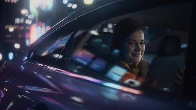 Stylish Female is Commuting Home in a Backseat of a Taxi at Night. Beautiful Woman Passenger Using Tablet Computer and Looking Out of Window while in Car in Urban City Street with Working Neon Signs.