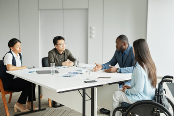 Diverse business team discussing project at meeting table in modern office with focus on young woman in wheelchair listening to colleagues
