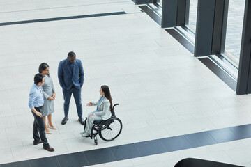 Graphic wide angle view at young businesswoman in wheelchair talking to diverse group of people in...