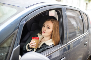 One girl with long hair is sitting in a black car, the view from the window. Holding a glass of coffee, thinking.