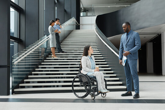 Wide angle view at diverse group of business people in office lobby focus on successful young woman in wheelchair talking to male colleague in foreground