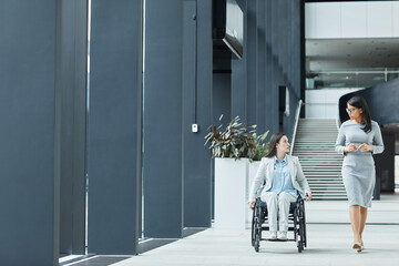 Full length portrait of young businesswoman in wheelchair talking to female colleague while moving towards camera in office lobby, copy space