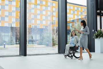 Full length portrait of young businesswoman in wheelchair talking to female colleague while moving...