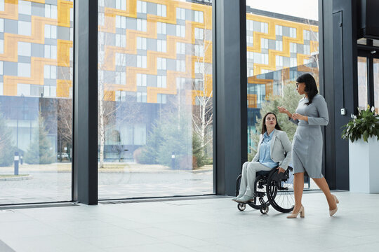 Full length portrait of young businesswoman in wheelchair talking to female colleague while moving against windows in office lobby, copy space