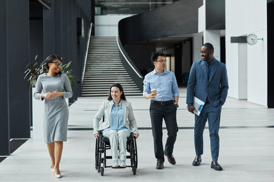Full length view at diverse group of business people chatting in graphic office lobby while moving towards camera, focus on smiling young woman in wheelchair