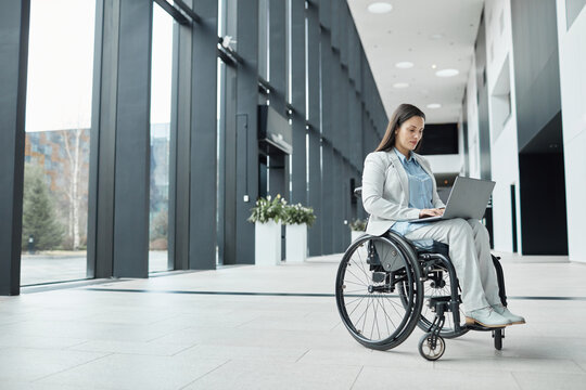 Full length portrait of successful businesswoman in wheelchair using laptop in office lobby, copy space