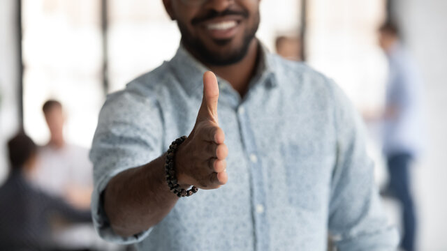 Happy mixed race employer or business owner welcoming for handshake and job. Black project leader giving hand for shake at camera, offering deal or partnership. Team working in background. Close up