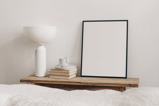 Cup of coffee on pile of books and modern marble geometric lamp. Vertical black picture frame mockup on vintage bench, table. White wall background. Scandinavian interior, bedroom. Selective focus.