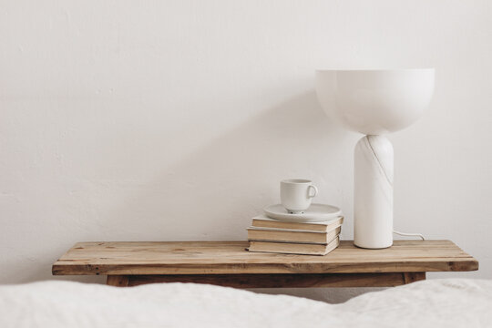 Cup of coffee on pile of books and modern marble geometric lamp. Vintage bench, table. White wall background. Scandinavian interior, bedroom. Selective focus. Blurred bedding. Empty copy space.