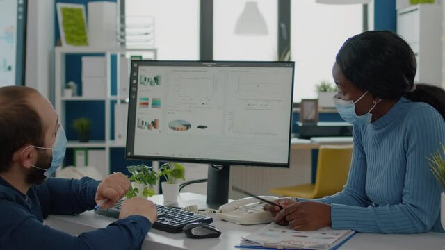 Handicapped paralysed invalid entretreneur with face mask explaning to black employee financial evolution of company, working together in new normal business office respecting social distance