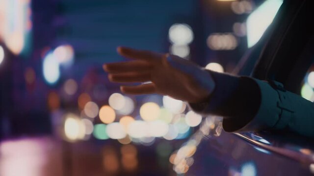 Female is Commuting Home in a Backseat of a Taxi at Night. Passenger Chilling and Holding Her Hand Outside of Window while in a Car in Urban City Street with Working Neon Signs. Cinematic Footage.