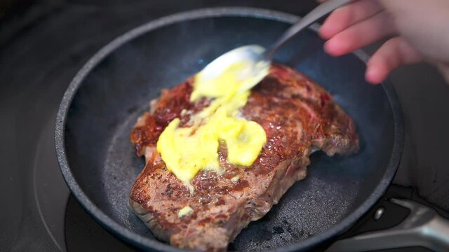 Macro closeup chef cook hand spreading butter on top of uncooked rare red grilled grass-fed ribeye rib eye meat steak on frying pan grill with pepper salt seasoning at home kitchen oven stove