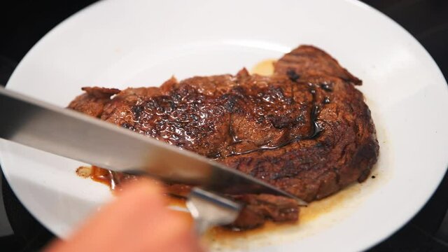 Macro closeup of person hands cutting with knife cooked rare medium red grilled grass-fed ribeye rib eye meat beef steak on white plate at home kitchen