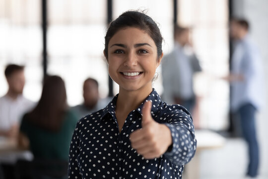 Photo portrait of happy Indian female business leader gesturing like, showing thumb up. Team working in background. Satisfied client woman gives approval positive feedback, recommends company service