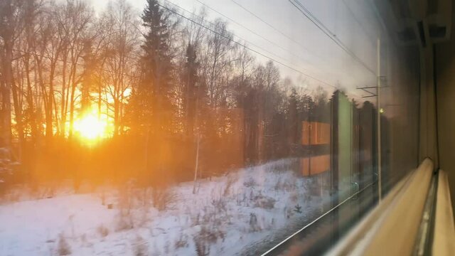 Setting sun flashes behind high bare and fir trees along power transmission lines in snowy valley view through driving train window