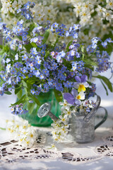 Obraz na płótnie Canvas A bouquet of spring blue forget-me-not flowers in a vase on the table, a tablecloth, bird cherry, pansies and a decorative watering can. Selective focus, blur, sunlight.