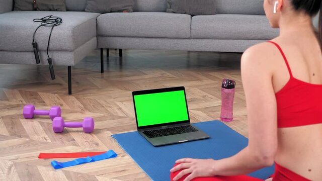 Green screen mock up chroma key monitor laptop: Fit woman in sportswear sits on yoga mat looks computer display, tells listen fitness trainer online video call webcam chat, distance sport course