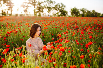 Fototapeta premium Beautiful young woman enjoys sitting among red poppy flowers on the field on the sunset. A girl feels harmony with nature and touches the plants. Love, peace and satisfaction concept.