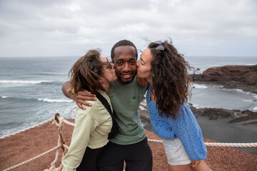 An African guy receives a kiss from two Caucasian girls during a vacation, a happy multiracial group is having fun.