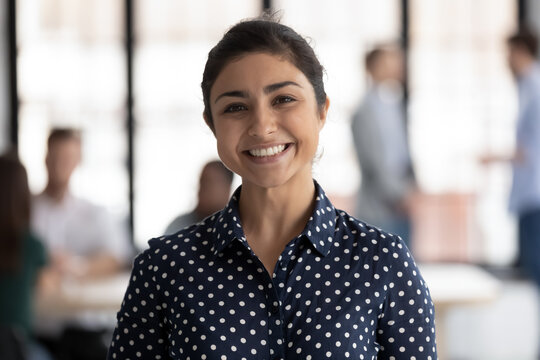 Corporate portrait of happy Indian female business team leader. Millennial employee looking at camera and smiling while group working in office behind her. Businesswoman, leadership concept. Head shot