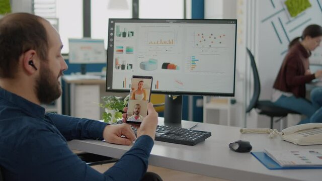 Disabled, invalid businessman in wheelchair discussing with friends on video call holding phone using headphones taking break sitting in business office. Diverse coworkers planning financial project