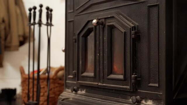 Metal fireplace with chimney and burning wood warming up a cabin or house. Fire burns inside a metal cast fire place with doors in cold winter or romantic night. Close up. Tools, coats in background