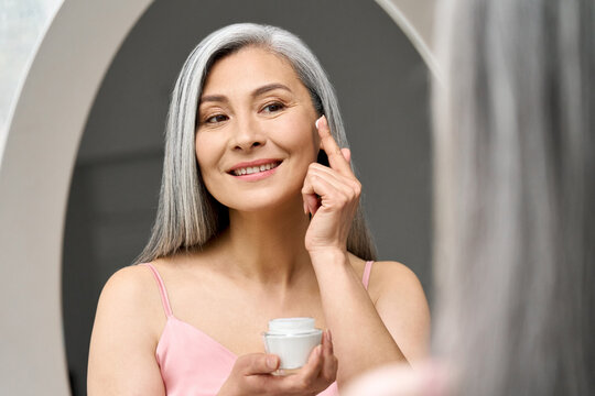 Mature beautiful happy middle aged asian woman, older grey haired lady looking at mirror touching face applying whitening antiaging anti wrinkle perfect skin care cream.