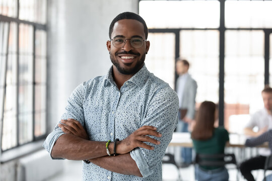 Portrait of happy African American small business owner posing with hands folded. Millennial black male team leader smiling, looking at camera, employees working in modern office behind. Head shot