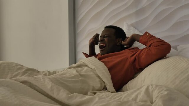 Young African-American man in pajamas awakes and gets out of bed to open curtains in stylish light bedroom in sunny morning