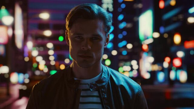 Portrait of Handsome Serious Man Turning and Looking at Camera, Standing in Night City with Bokeh Neon Street Lights in Background. Confident Young Man with Stylish Hair. Close-up Shot