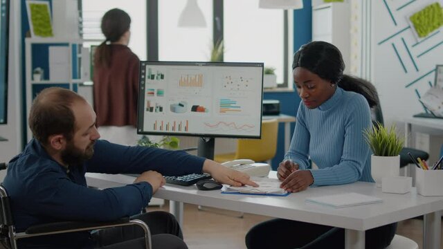 Diverse team working together with financial graphs discussing solution for project deadline in strat up business office, black woman and paralysed coworker in wheelchair comparing informations