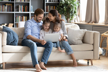 Fototapeta premium Happy young Caucasian family renters with two small daughters relax in living room with modern laptop. Smiling parents with little girls children rest on sofa at home using computer together.