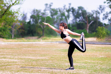 Woman doing yoga exercises on the lawn. Person stands on one leg, extending the right arm forward, the left arm holding the left leg bent back. Concept of exercise among nature.