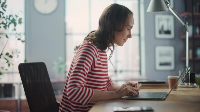 Young Woman Using Laptop at Home for Remote Work. Beautiful Young Brunette Girl Sitting at Her Desk Uses Internet, Doing Productive Distant Work. Side View Medium Arc Shot